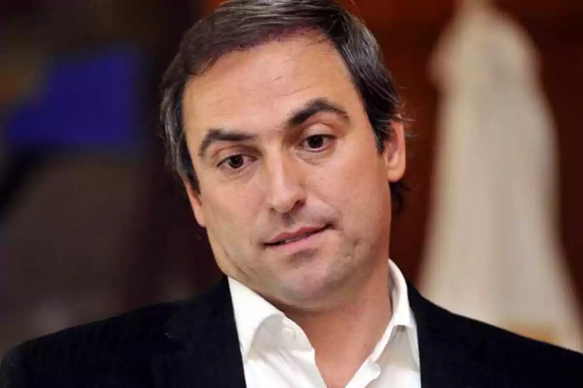 Short-haired, gray-haired man with a serious expression wears a white jersey and dark jacket in a blurred indoor setting. Short-haired, gray-haired man with a serious expression wears a white jersey and dark jacket in a blurred indoor setting.