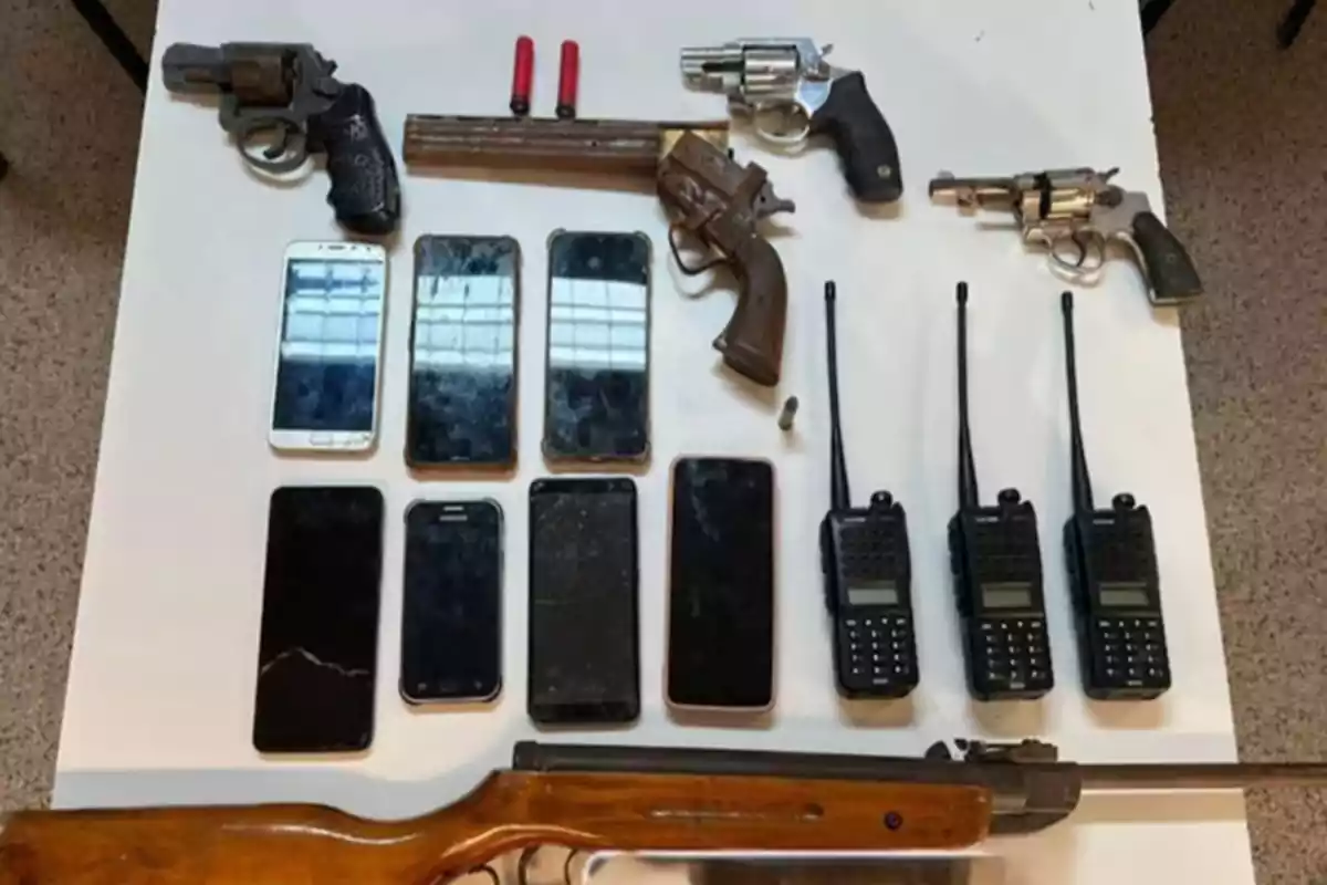 Sobre una mesa blanca hay varias armas de fuego, teléfonos móviles, radios portátiles y cartuchos de munición.