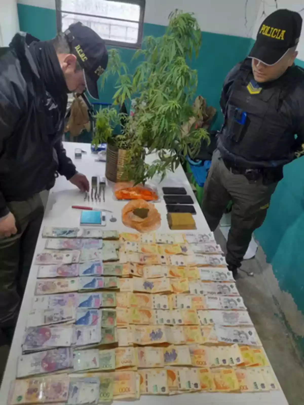 Dos policías observan una mesa con billetes, plantas de marihuana, teléfonos, balanza y otros objetos incautados