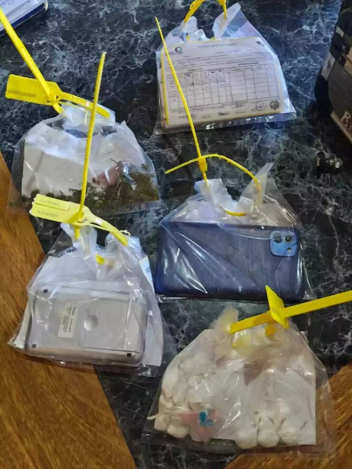 Cinco bolsas de plástico selladas con cintillos amarillos contienen diferentes objetos como documentos, una cartera, una báscula, hierba y varias bolsas pequeñas sobre una mesa de mármol.