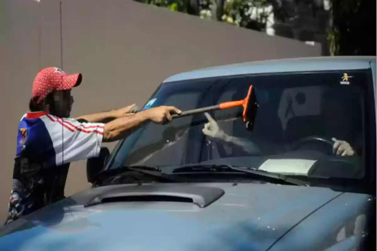Un hombre con gorra y camiseta deportiva limpia el parabrisas de un automóvil en la calle mientras el conductor está dentro del vehículo