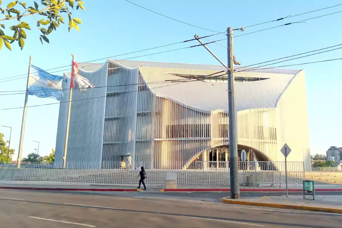 Edificio moderno de fachada blanca con formas onduladas y columnas verticales, dos banderas ondeando al frente y una persona caminando por la acera en un día soleado Edificio moderno de fachada blanca con formas onduladas y columnas verticales, dos banderas ondeando al frente y una persona caminando por la acera en un día soleado