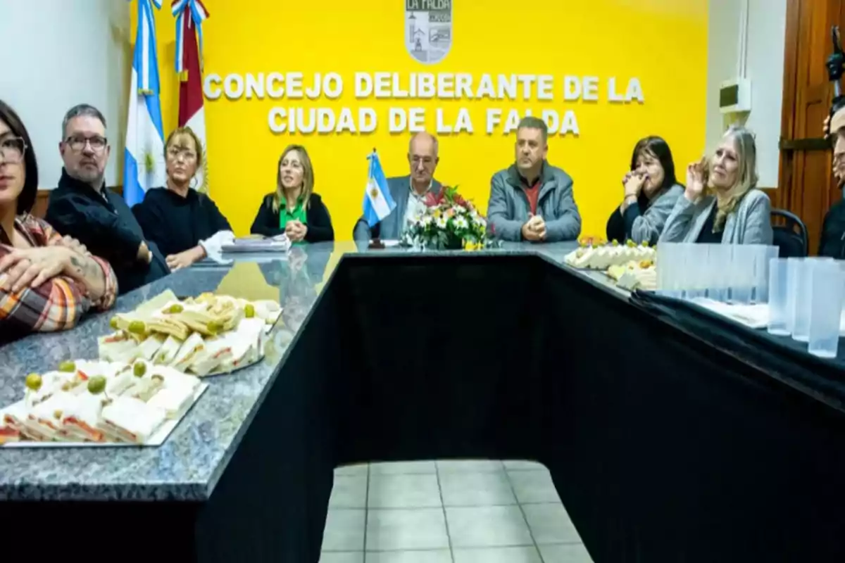 Reunión de varias personas sentadas alrededor de una mesa en una sala con paredes amarillas, al fondo se lee Concejo Deliberante de la Ciudad de La Falda y sobre la mesa hay bandejas con sándwiches y vasos de plástico.