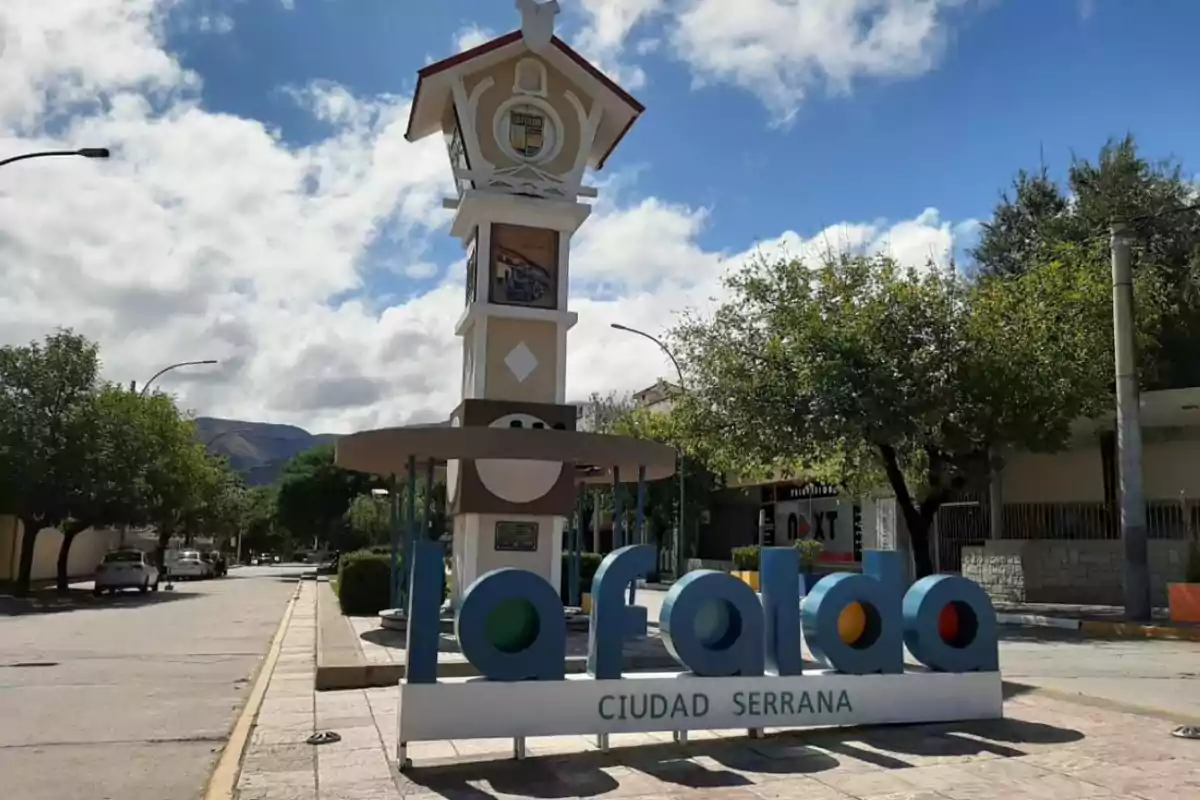 Cartel colorido de bienvenida a la ciudad de La Falda con un reloj de torre al fondo en una calle arbolada y cielo parcialmente nublado