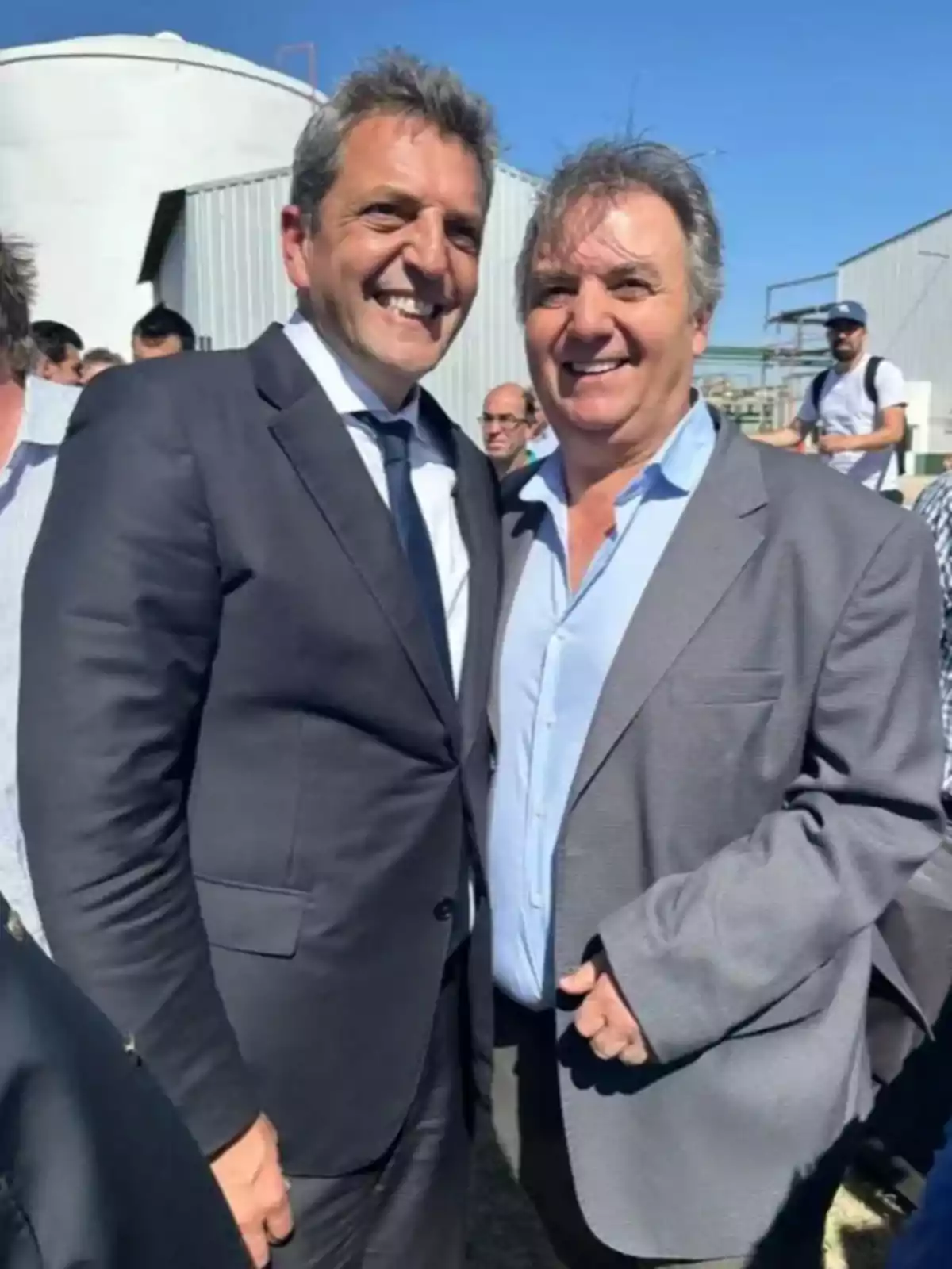 Two men smile and pose together outdoors, both dressed in suits and ties, with a background of blue sky and industrial structures. Two men smile and pose together outdoors, both dressed in suits and ties, with a background of blue sky and industrial structures.
