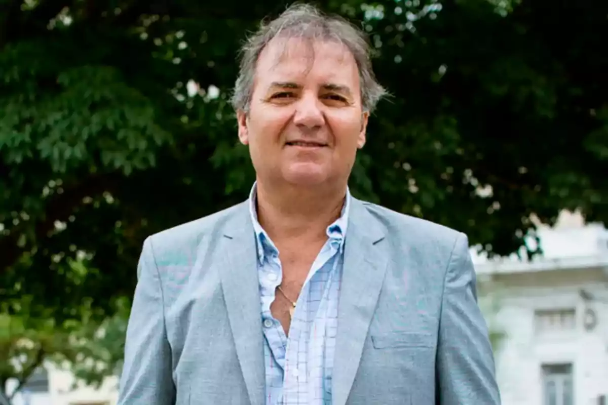 Middle-aged man with gray hair wearing a gray jacket and light blue jersey posing outdoors in front of trees