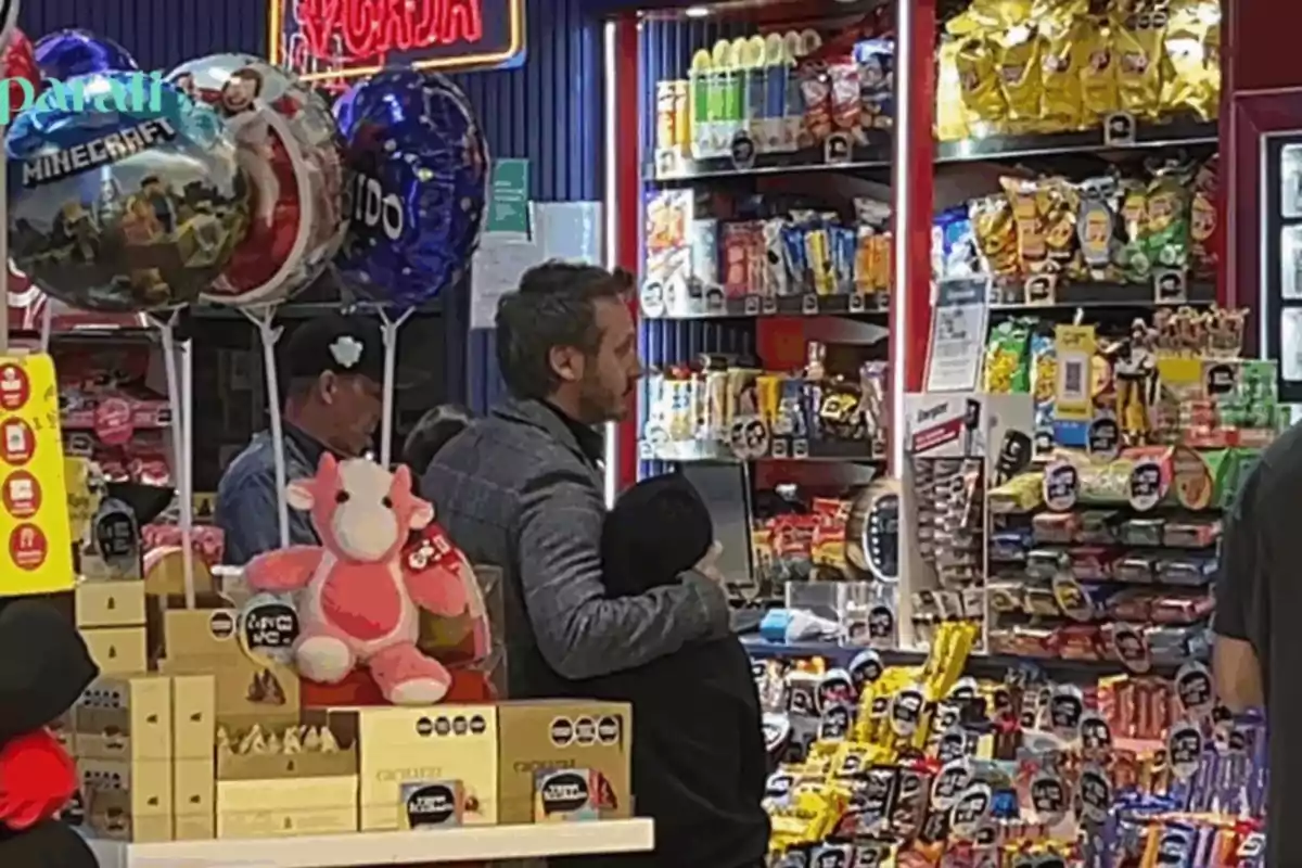 A man hugs a boy while they wait in line at a candy and snack shop, surrounded by balloons and stuffed animals. A man hugs a boy while they wait in line at a candy and snack shop, surrounded by balloons and stuffed animals.