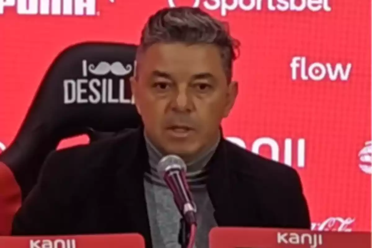 Hombre de cabello corto y canoso hablando frente a un micrófono en una conferencia de prensa con fondo rojo y logotipos de marcas.