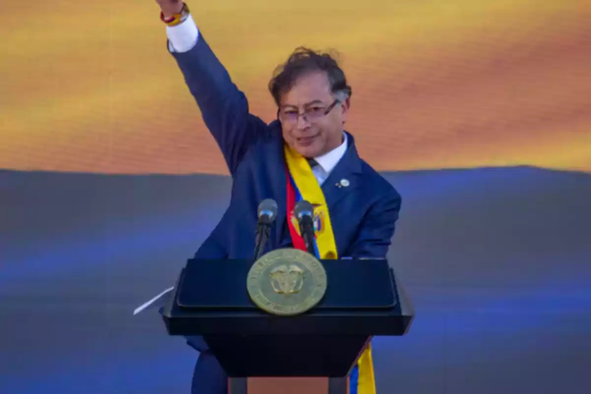 Man with a blue, yellow, and red presidential sash raising his right arm behind a podium with microphones and the national coat of arms Man with a blue, yellow, and red presidential sash raising his right arm behind a podium with microphones and the national coat of arms