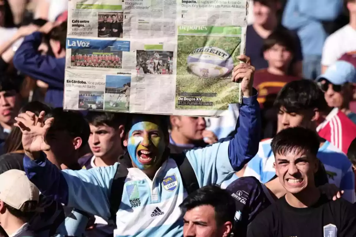 Fan with a sky blue and yellow painted face holds a newspaper above his head while celebrating in a crowd during a rugby match. Fan with a sky blue and yellow painted face holds a newspaper above his head while celebrating in a crowd during a rugby match.