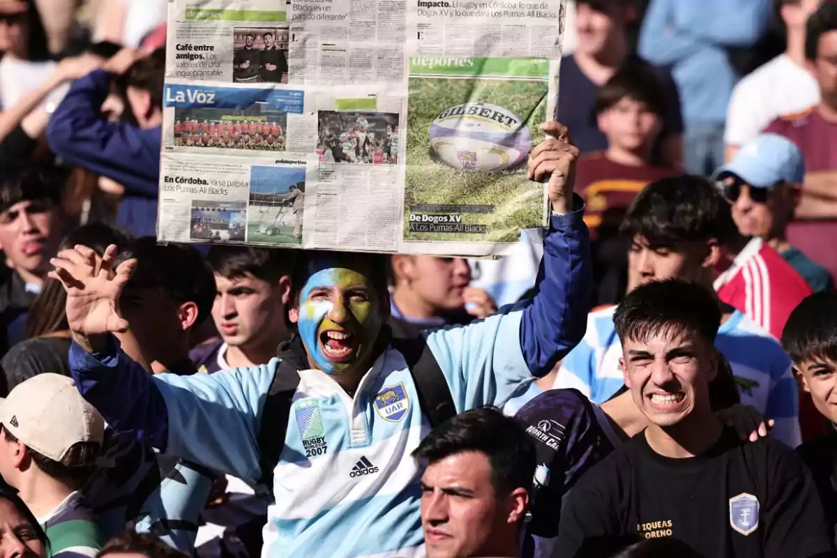 Aficionados de rugby animando en las gradas, uno de ellos con la cara pintada y sosteniendo un periódico sobre su cabeza para protegerse del sol