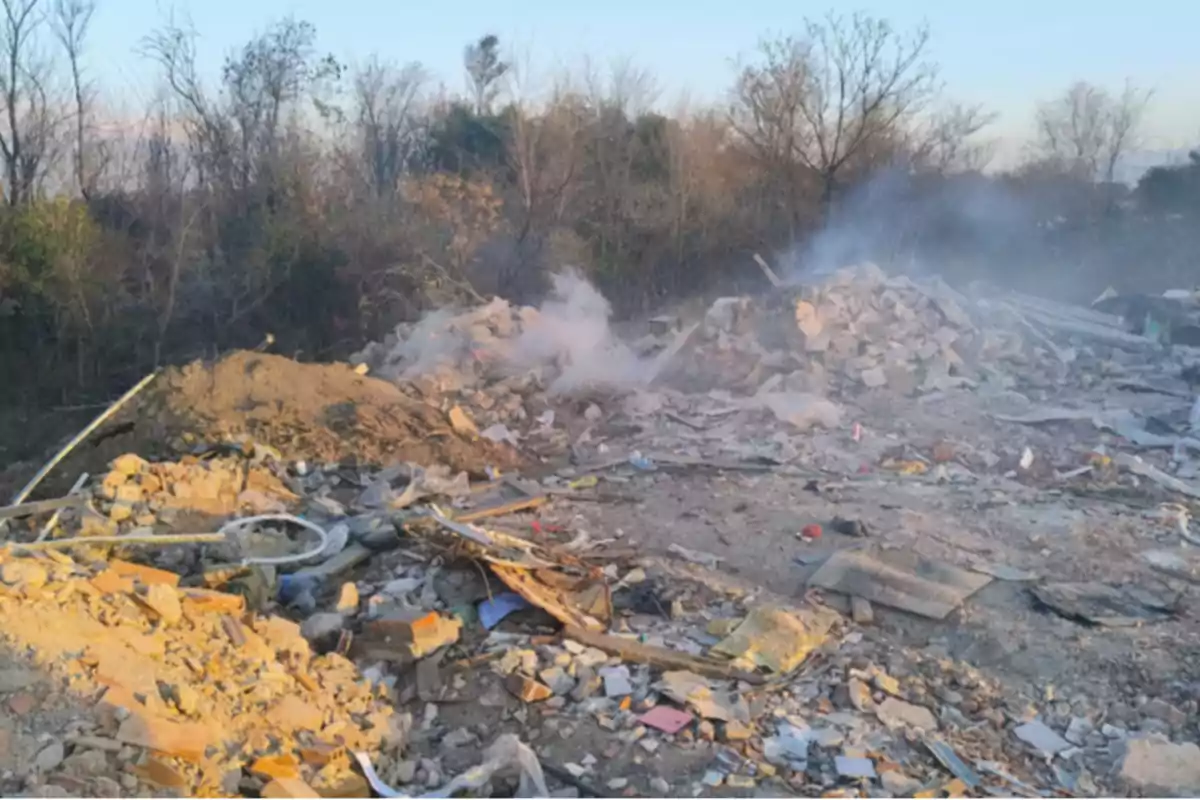 Debris and trash scattered on an open field with smoke rising from one of the piles and trees in the background. Debris and trash scattered on an open field with smoke rising from one of the piles and trees in the background.