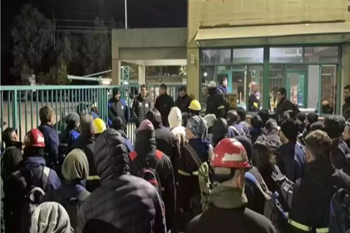 Un grupo numeroso de personas con cascos y ropa de trabajo se reúne frente a una entrada de edificio durante la noche