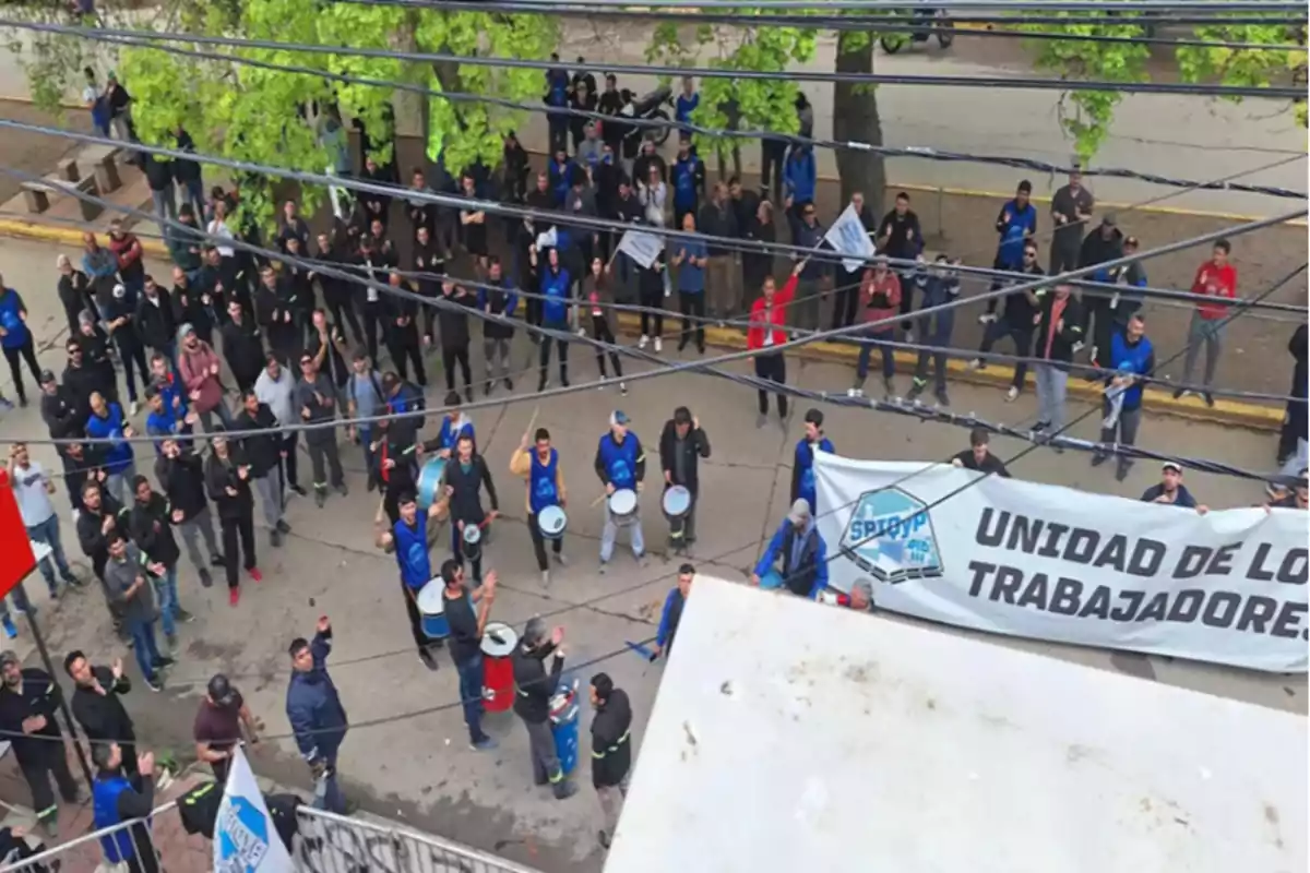 Manifestación de trabajadores en la calle con pancarta y tambores, vista desde arriba, rodeados de árboles y cables eléctricos