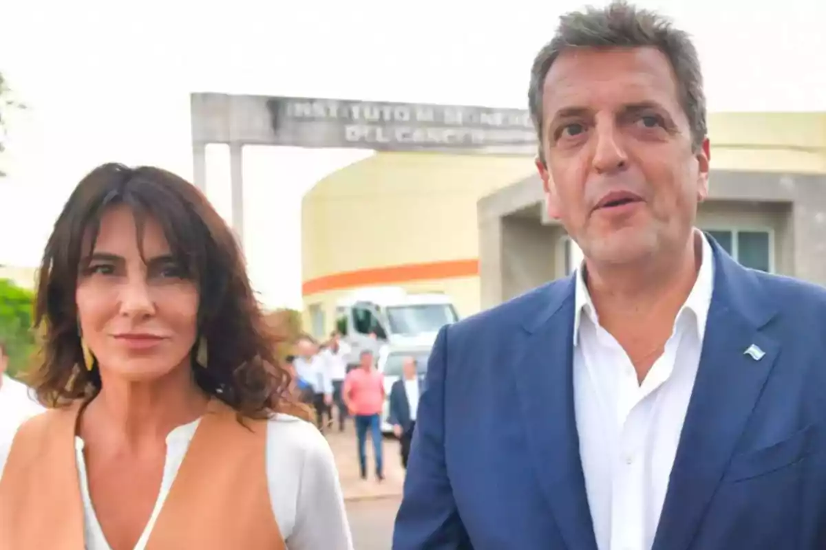 Una mujer y un hombre vestidos de manera formal están al aire libre frente a un edificio con un cartel que dice Instituto del Cáncer, hay más personas y una ambulancia en el fondo.