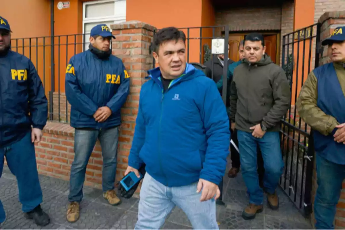 Un grupo de hombres, algunos con chaquetas y gorras de la PFA, están de pie frente a una casa de ladrillos y portón negro, mientras uno de ellos camina hacia adelante sosteniendo un teléfono móvil.