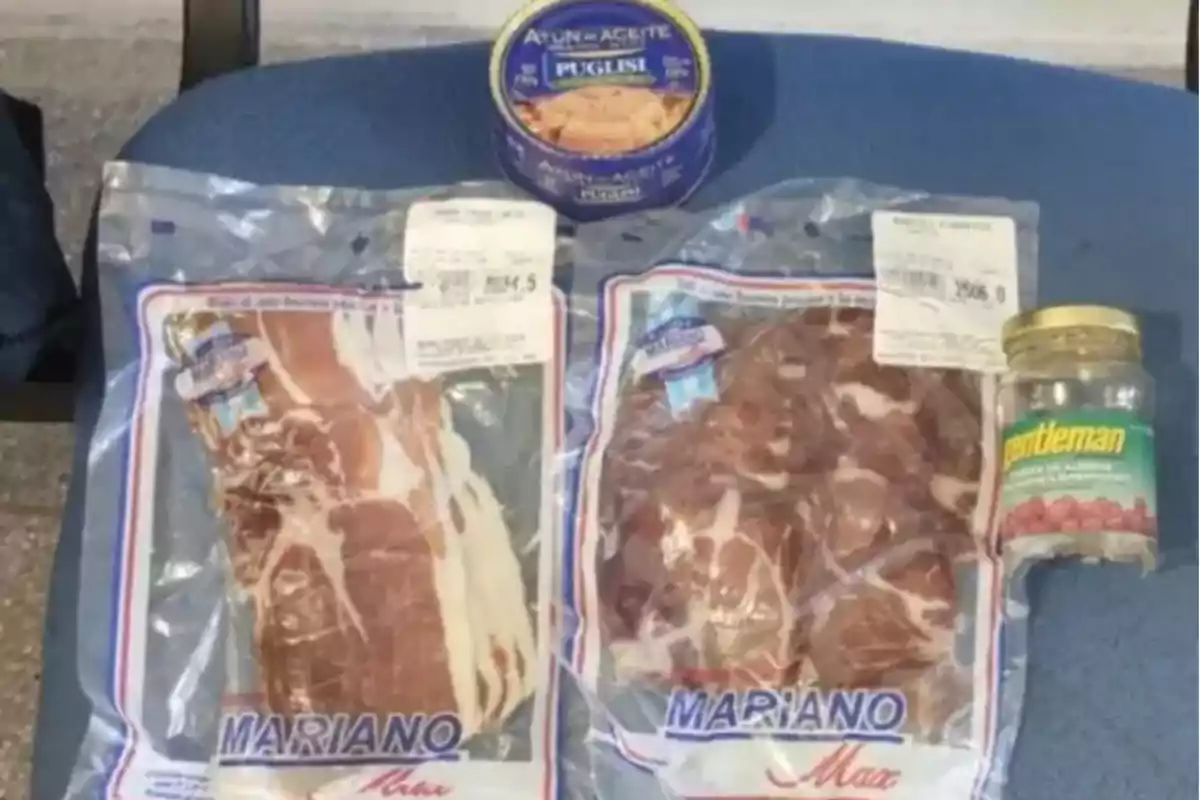 Paquetes de carne embutida, una lata de atún y un frasco de aceitunas sobre una superficie azul