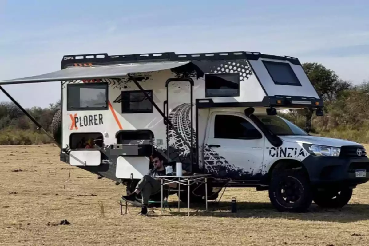 Camper-type pickup truck equipped for camping in the open field with a person sitting under the awning next to a folding table Camper-type pickup truck equipped for camping in the open field with a person sitting under the awning next to a folding table