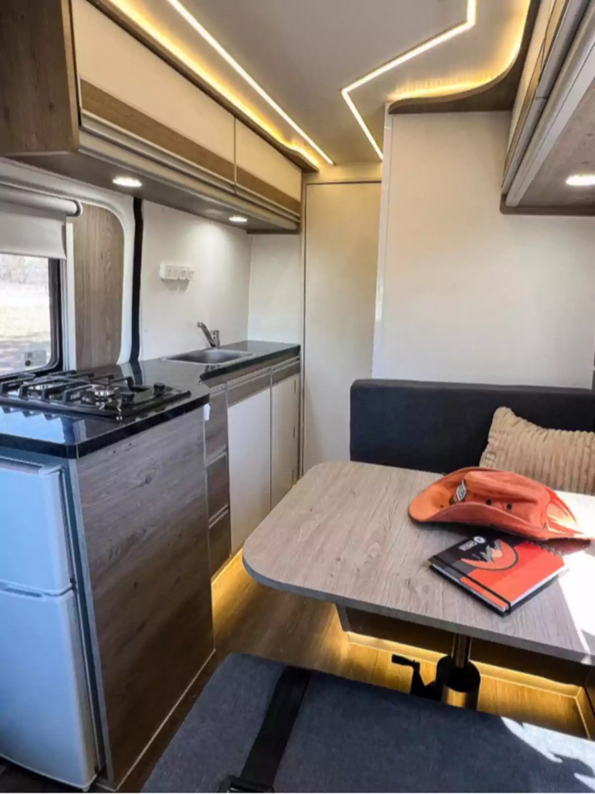 Modern interior of a motorhome with kitchen, dining table, notebook and orange hat on the table, warm LED lighting, and light wood finishes Modern interior of a motorhome with kitchen, dining table, notebook and orange hat on the table, warm LED lighting, and light wood finishes