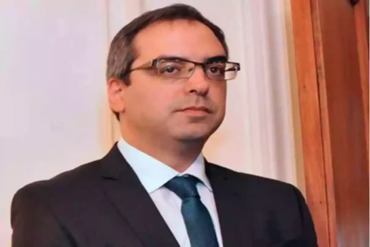 Hombre de cabello corto y gafas vistiendo traje oscuro con corbata azul en un entorno interior
