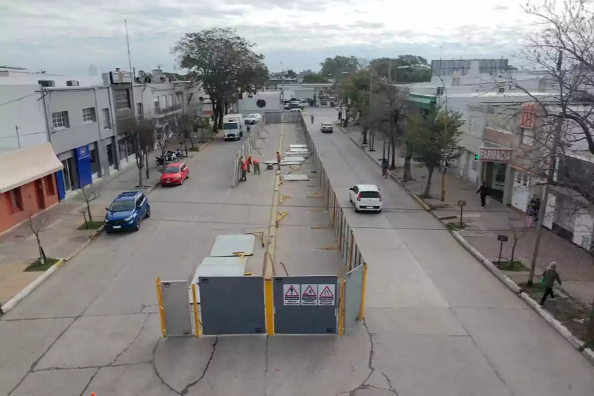 Calle urbana bloqueada por vallas metálicas con trabajadores usando chalecos naranjas y algunos autos estacionados y circulando