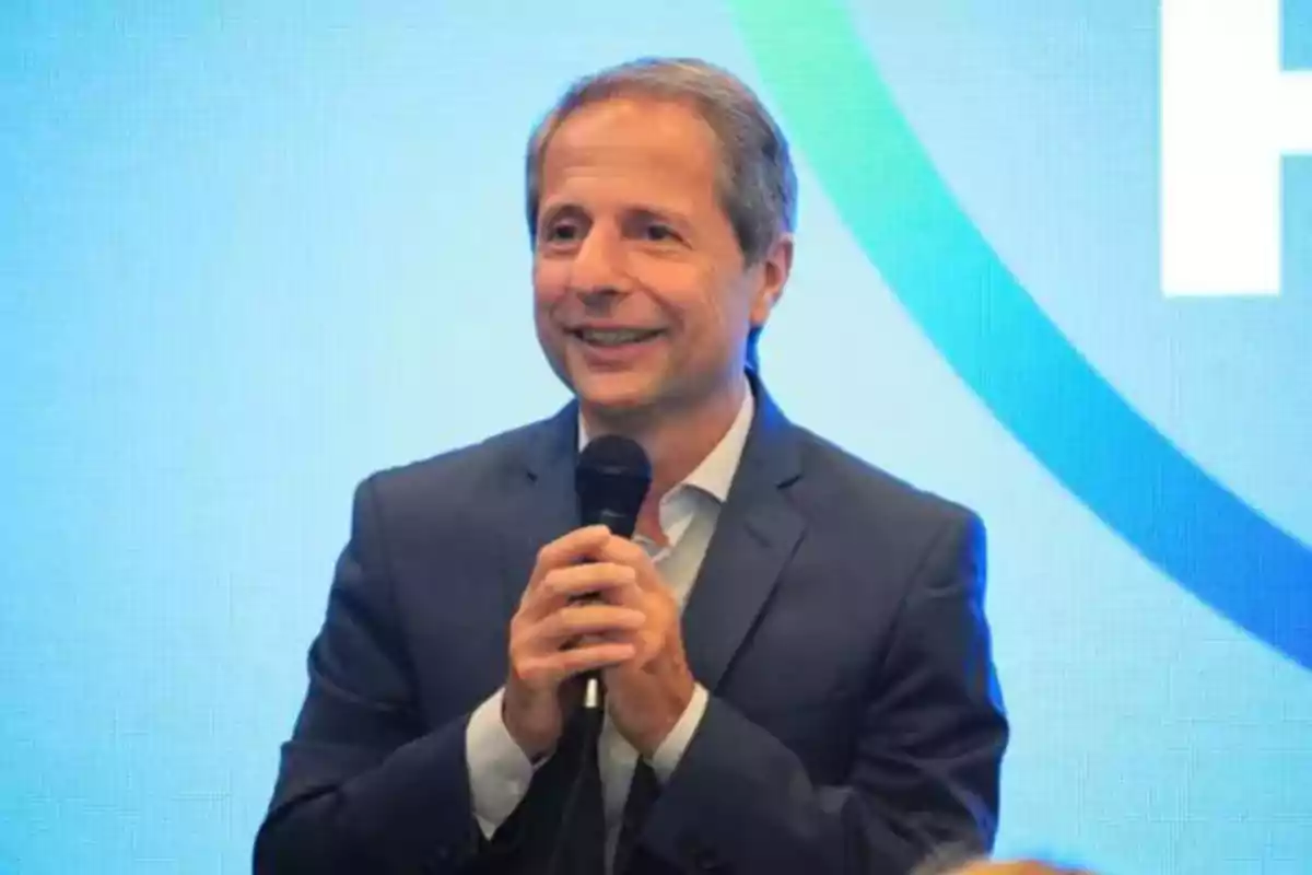 Hombre de traje sosteniendo un micrófono y sonriendo frente a un fondo azul claro