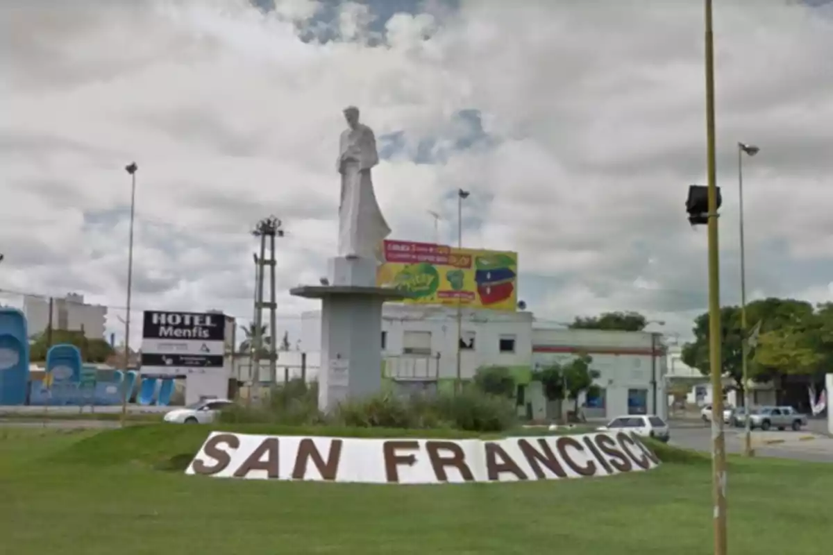 Rotonda con una estatua blanca de un santo sobre un pedestal en una zona urbana con césped y un letrero que dice San Francisco