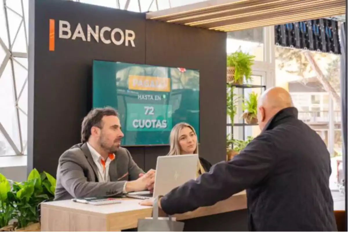 Tres personas interactúan en un stand de atención de Bancor con plantas decorativas y una pantalla que promociona pagos en cuotas