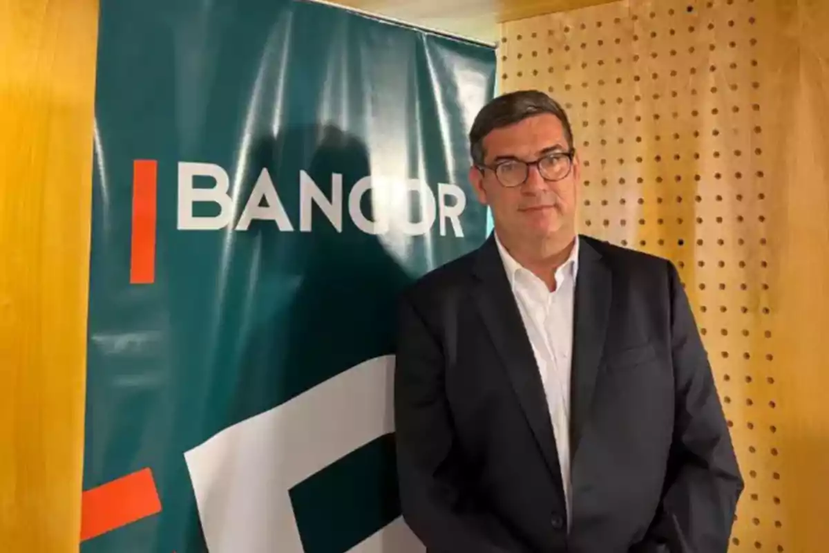 Hombre con gafas y traje oscuro posando junto a un cartel de Bancor en un ambiente interior con paredes de madera. Hombre con gafas y traje oscuro posando junto a un cartel de Bancor en un ambiente interior con paredes de madera.