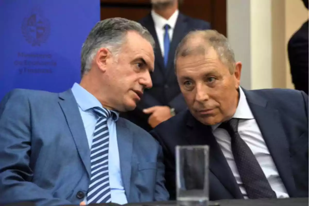Dos hombres de traje conversan en una reunión formal con un vaso de agua sobre la mesa y un cartel del Ministerio de Economía y Finanzas al fondo