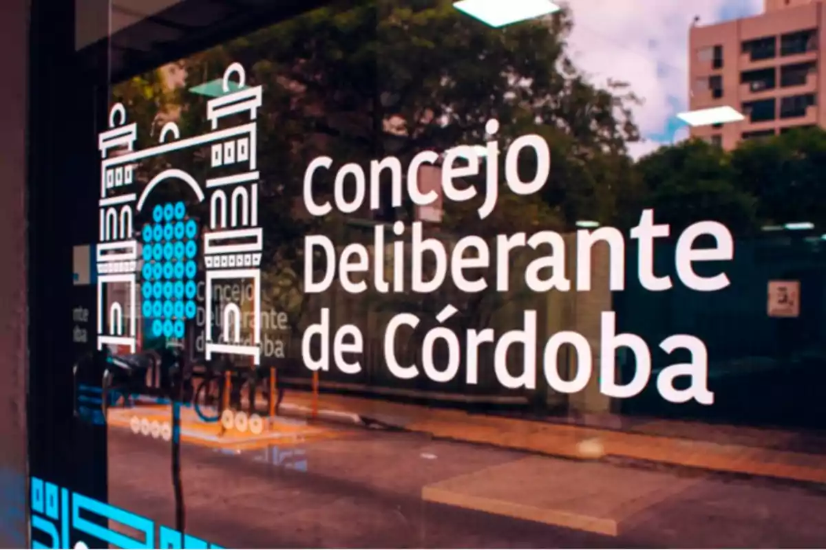 Puerta de vidrio con el logo y el nombre Concejo Deliberante de Córdoba reflejando la calle y árboles al fondo Puerta de vidrio con el logo y el nombre Concejo Deliberante de Córdoba reflejando la calle y árboles al fondo
