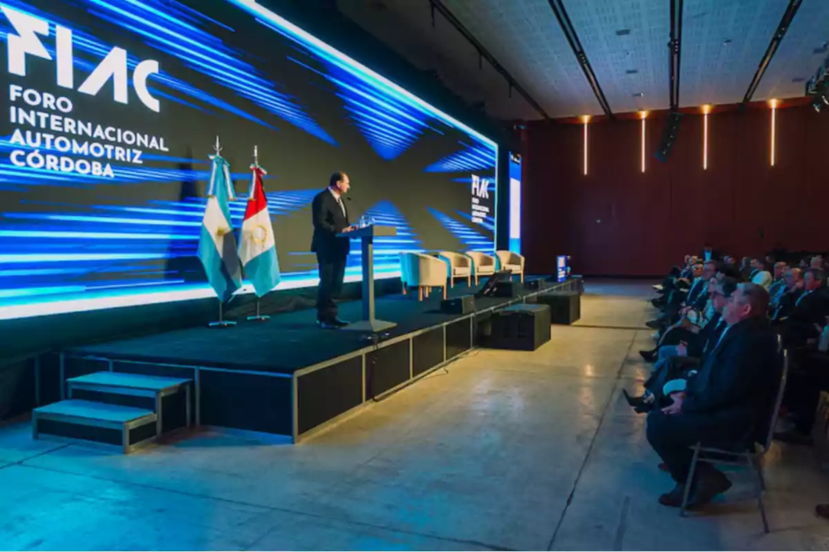 A man in a suit speaks on stage during an automotive event in Córdoba, with an audience seated and two flags next to the podium.