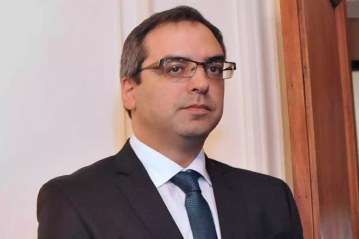 Hombre de cabello corto y gafas vistiendo traje oscuro y corbata azul en un entorno interior