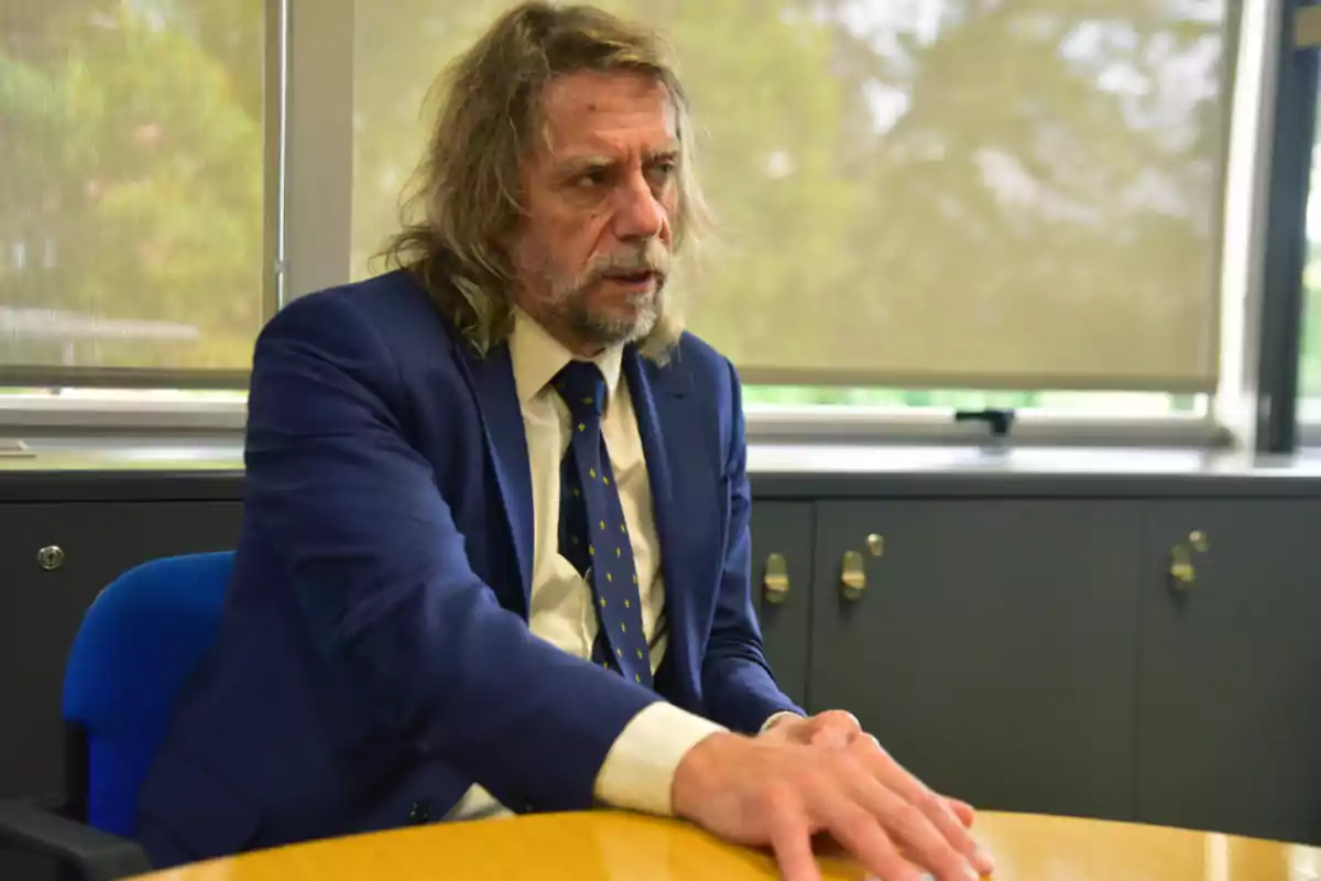 Long-haired, gray-haired man with a beard, wearing a blue suit and tie, sitting in an office next to a round table Long-haired, gray-haired man with a beard, wearing a blue suit and tie, sitting in an office next to a round table