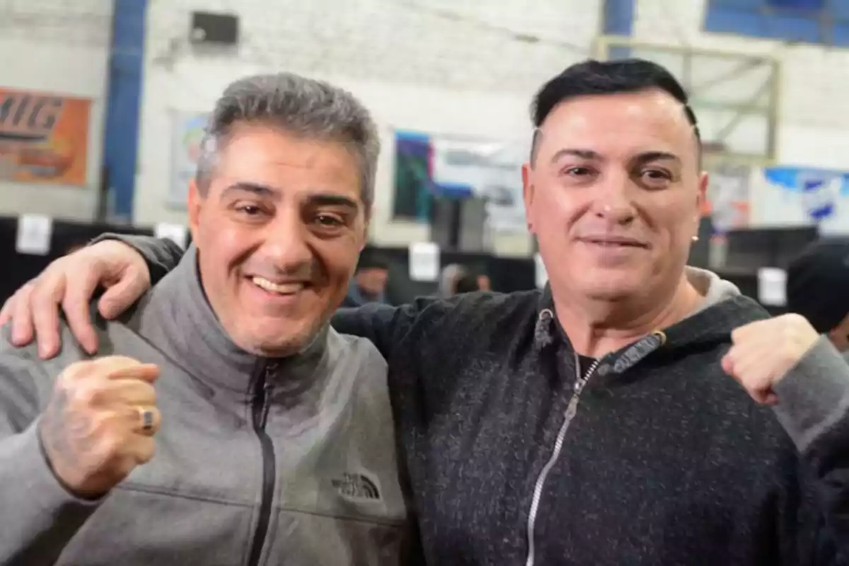 Two men smile and pose together indoors, both wearing sportswear and displaying a sense of camaraderie.