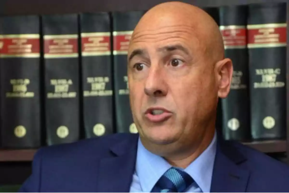 Bald man in a blue suit speaking in front of a shelf of law books