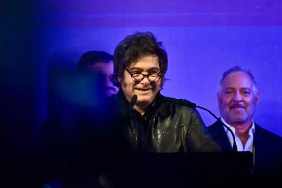 Man with glasses and a black leather jacket speaking into a microphone on a blue-lit stage with people in the background