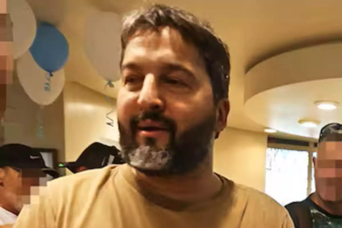 Hombre de barba y cabello oscuro en un ambiente interior con globos y personas al fondo