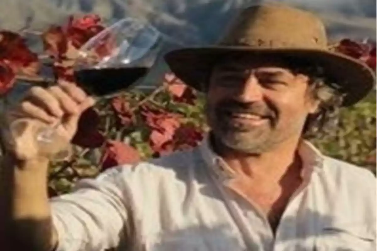 Hombre sonriente con sombrero sostiene una copa de vino tinto en un viñedo con hojas rojas al fondo Hombre sonriente con sombrero sostiene una copa de vino tinto en un viñedo con hojas rojas al fondo