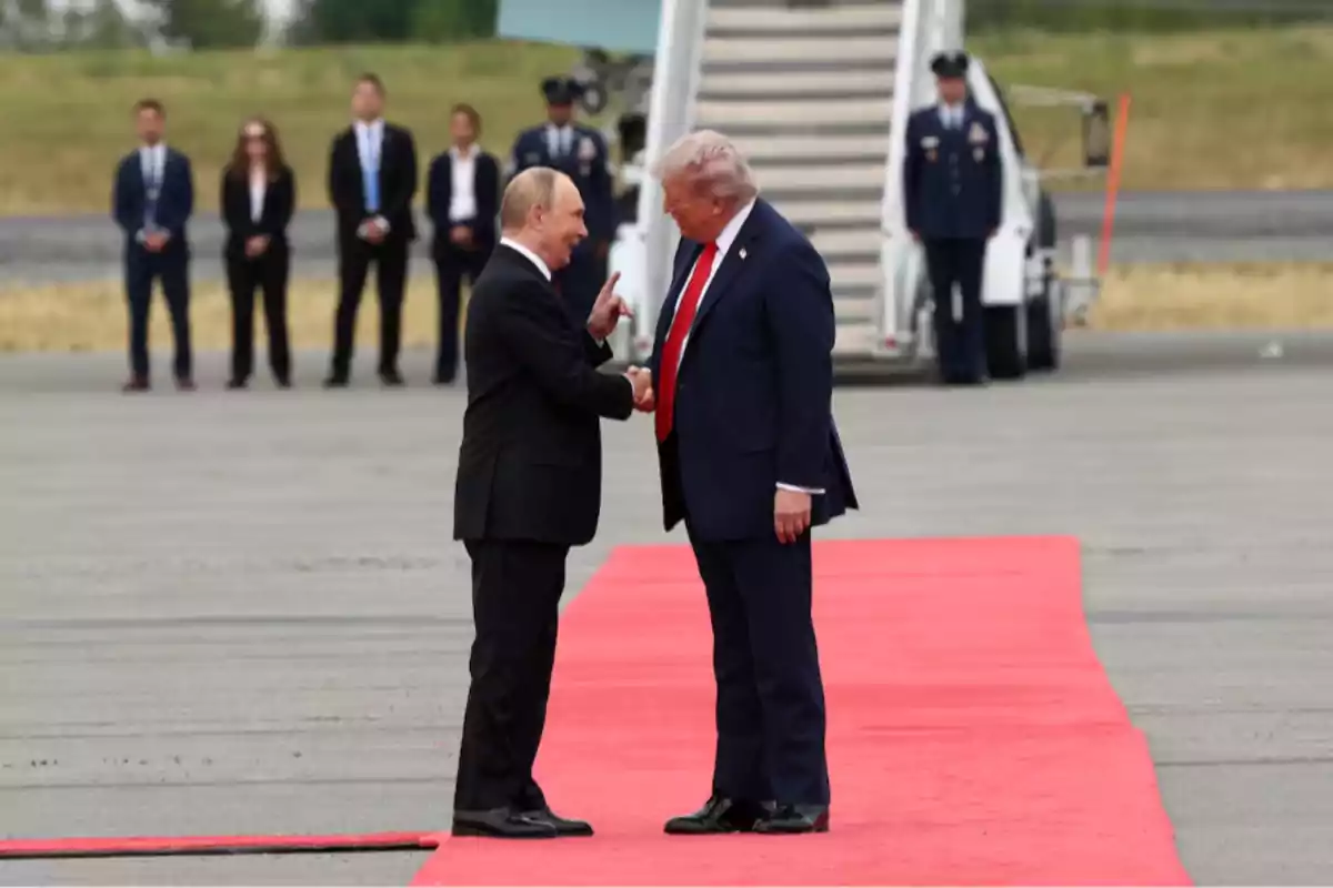 Two men in suits shake hands on a red carpet on an airport runway while several people watch in the background. Two men in suits shake hands on a red carpet on an airport runway while several people watch in the background.