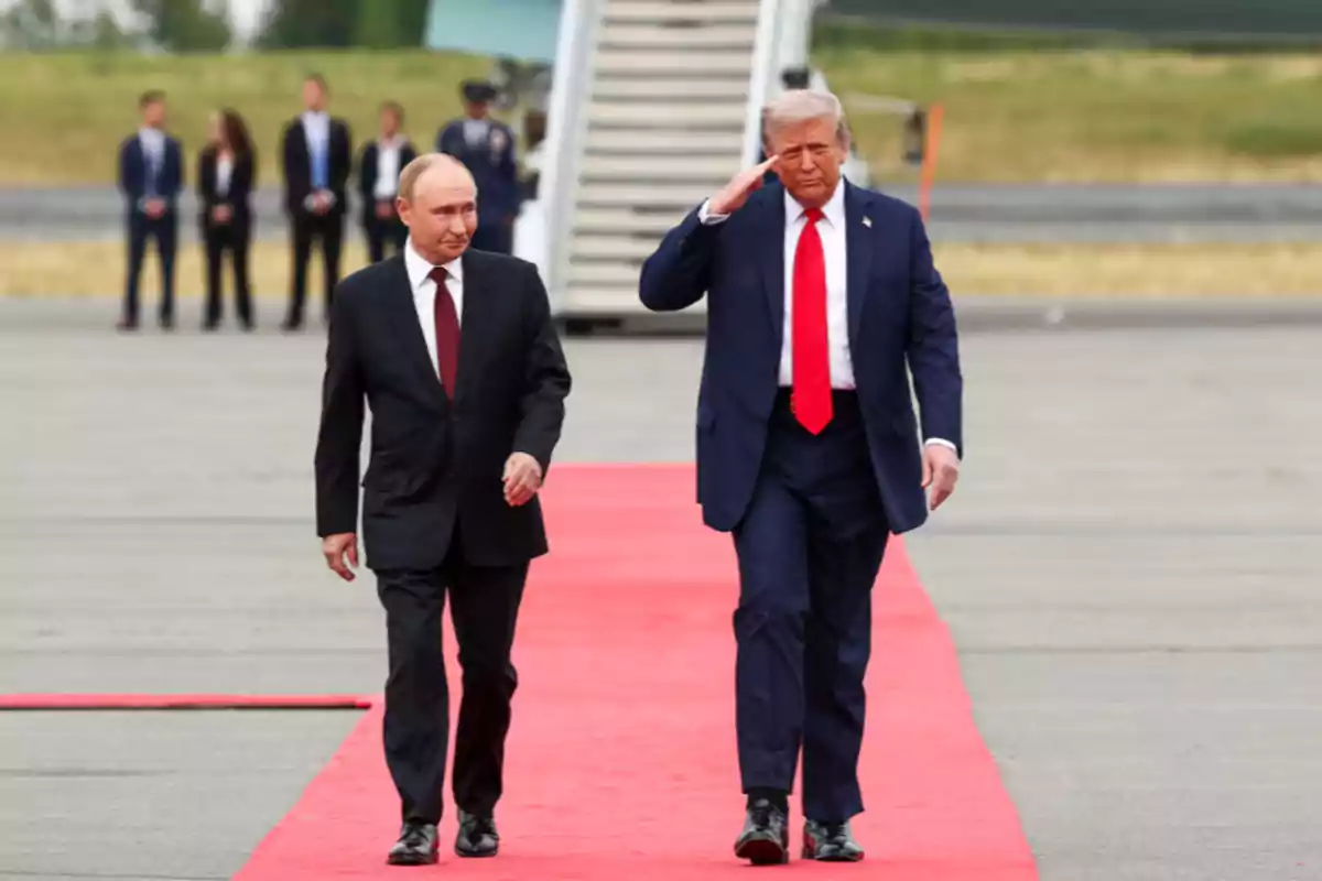 Two men in dark suits walk on a red carpet on an airport runway while one of them salutes with his hand on his forehead, and several people can be seen standing in the background. Two men in dark suits walk on a red carpet on an airport runway while one of them salutes with his hand on his forehead, and several people can be seen standing in the background.