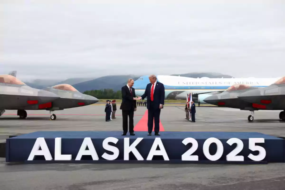 Two men in suits shake hands on a platform that says ALASKA 2025 on an airport runway with military planes and a presidential plane in the background.