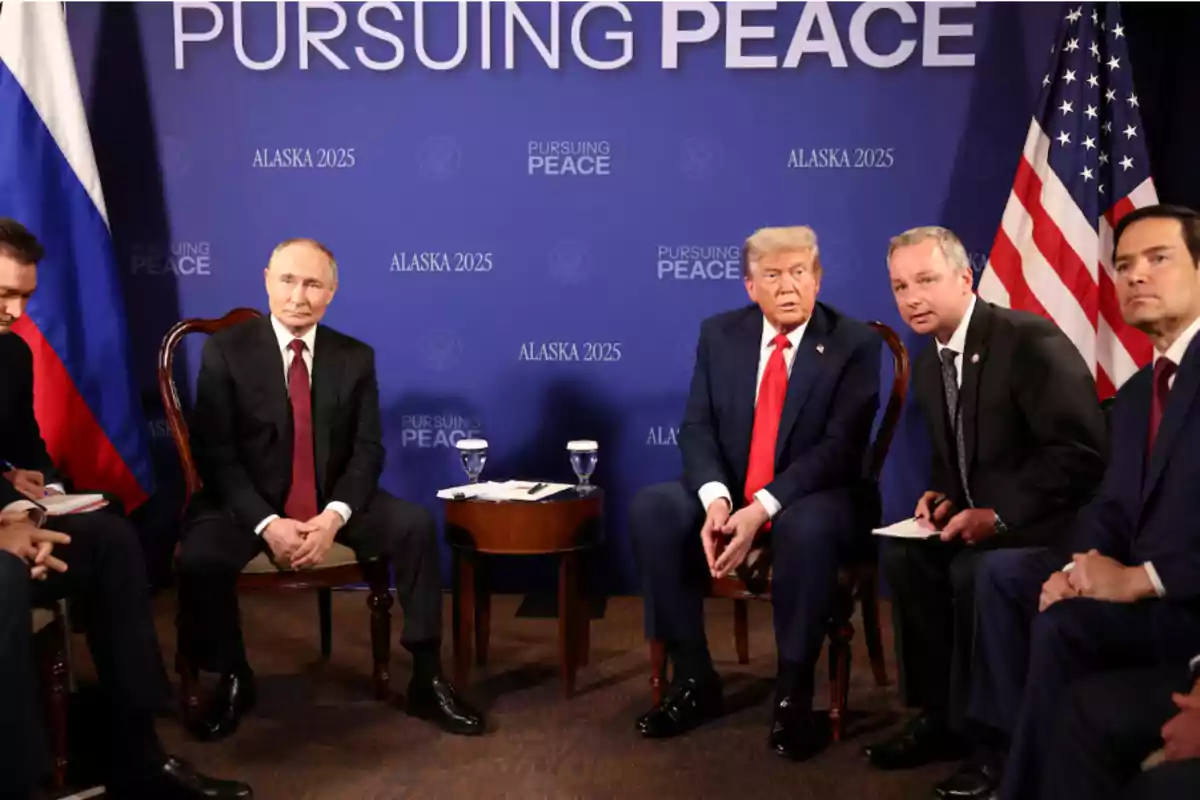 Five men in formal suits seated at a meeting in front of a blue background with the words Five men in formal suits seated at a meeting in front of a blue background with the words