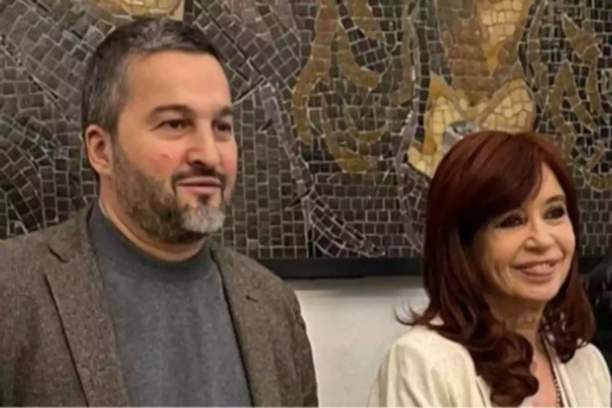 Two people pose smiling in front of a mosaic mural in dark tones. Two people pose smiling in front of a mosaic mural in dark tones.