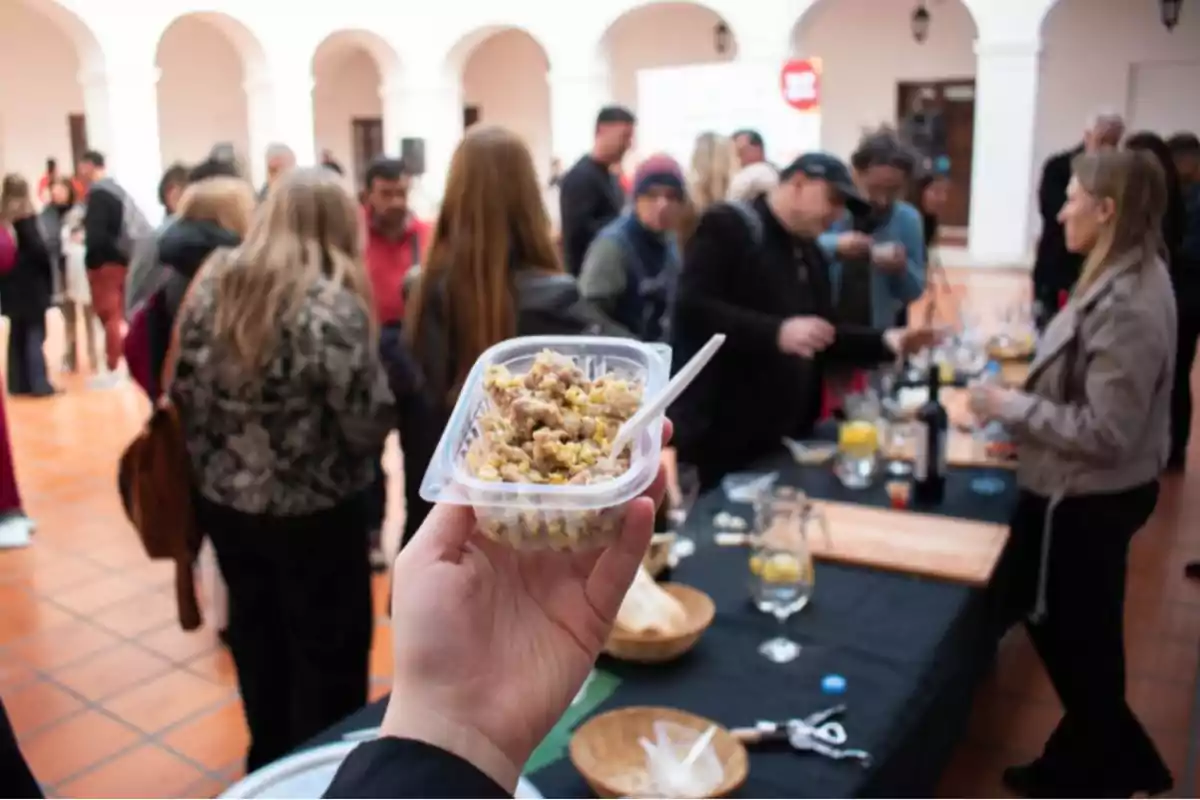 Persona sosteniendo un recipiente de comida con un tenedor de plástico en primer plano mientras varias personas conversan y degustan alimentos en una mesa durante un evento social en un espacio interior con arcos blancos.