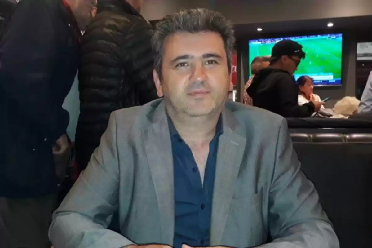 Hombre de cabello canoso y barba incipiente sentado en un restaurante con varias personas al fondo y una pantalla de televisión mostrando un partido de fútbol. Hombre de cabello canoso y barba incipiente sentado en un restaurante con varias personas al fondo y una pantalla de televisión mostrando un partido de fútbol.