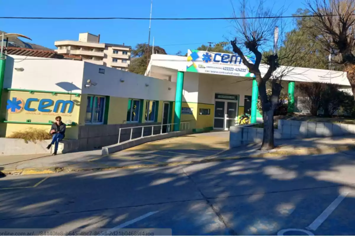 Fachada de un centro de emergencias médicas con una persona sentada en el borde de la vereda y árboles sin hojas en la acera Fachada de un centro de emergencias médicas con una persona sentada en el borde de la vereda y árboles sin hojas en la acera