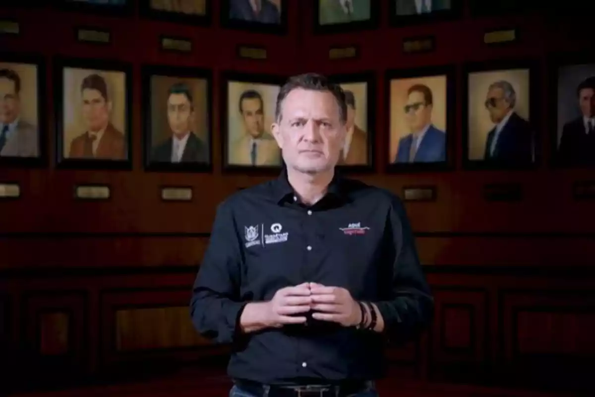 Man standing with a black jersey in front of a wall with framed portraits of various people