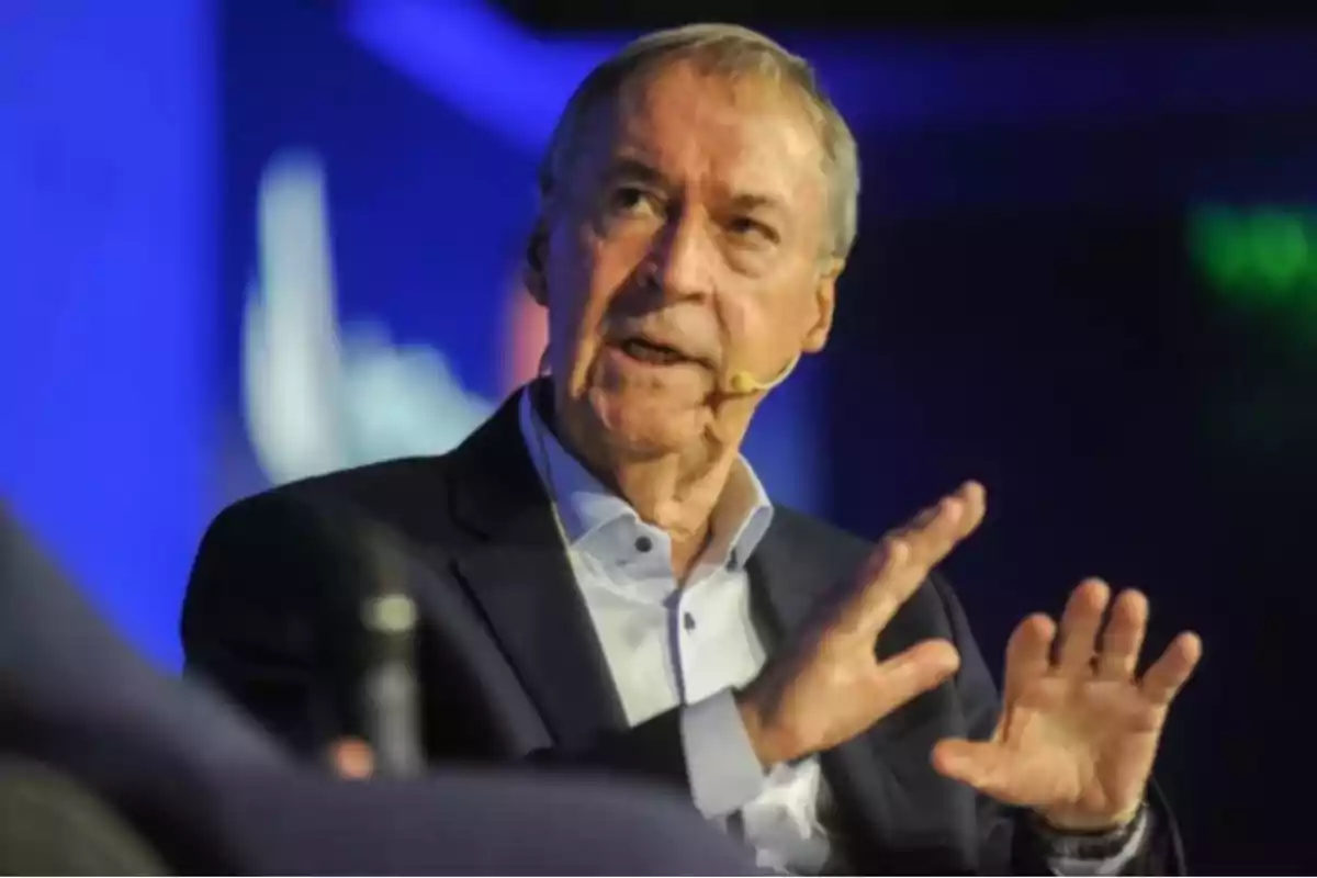 Older man with a headset microphone speaking and gesturing at an event with a blurred blue background Older man with a headset microphone speaking and gesturing at an event with a blurred blue background