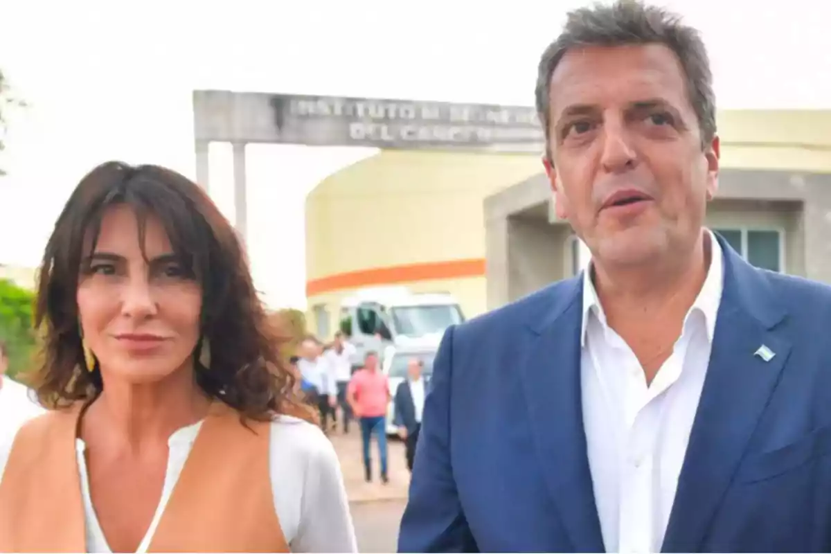 A woman and a man standing outdoors in front of a building with people and a vehicle in the background. A woman and a man standing outdoors in front of a building with people and a vehicle in the background.